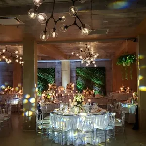Dans cette image de la salle de réception Plaza Volare, un espace élégant et bien éclairé est présenté pour un mariage. On y voit des tables rondes dressées avec des nappes blanches, des chaises transparentes et des centres de table floraux raffinés. Le plafond est orné de luminaires modernes, tandis que des murs végétaux apportent une touche naturelle et sophistiquée à l'ensemble. L'ambiance chaleureuse et accueillante de la salle crée un cadre parfait pour célébrer un mariage mémorable au Canada.