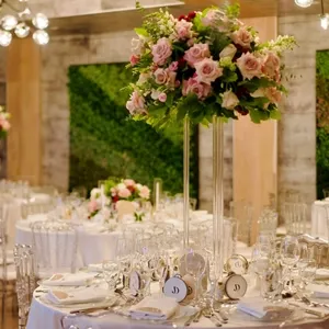 La salle de réception Plaza Volare est magnifiquement décorée pour un mariage, avec des tables élégantes ornées de nappes blanches et de vaisselle raffinée. Un grand arrangement floral de roses roses et de feuillage vert est placé au centre de chaque table, donnant une touche de romantisme et de sophistication. En arrière-plan, un mur végétalisé apporte une ambiance naturelle et luxueuse à l'espace. Les chaises transparentes ajoutent une touche moderne à l'ensemble. Cette image illustre parfaitement le cadre élégant et accueillant que la salle Plaza Volare offre pour célébrer un mariage mémorable au Canada.