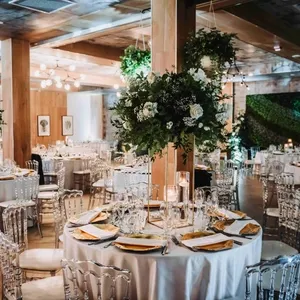 La salle de réception Plaza Volare est magnifiquement décorée pour un mariage élégant. Des tables rondes recouvertes de nappes blanches sont soigneusement dressées avec de la vaisselle raffinée et des serviettes dorées. De magnifiques compositions de fleurs blanches et vertes ornent le centre des tables, ajoutant une touche de sophistication. Les chaises transparentes modernes entourant les tables complètent l'ensemble, tandis que de grands piliers en bois et un éclairage chaleureux créent une ambiance accueillante. L'arrière-plan présente un mur végétal, apportant une ambiance naturelle et élégante à l'espace.
