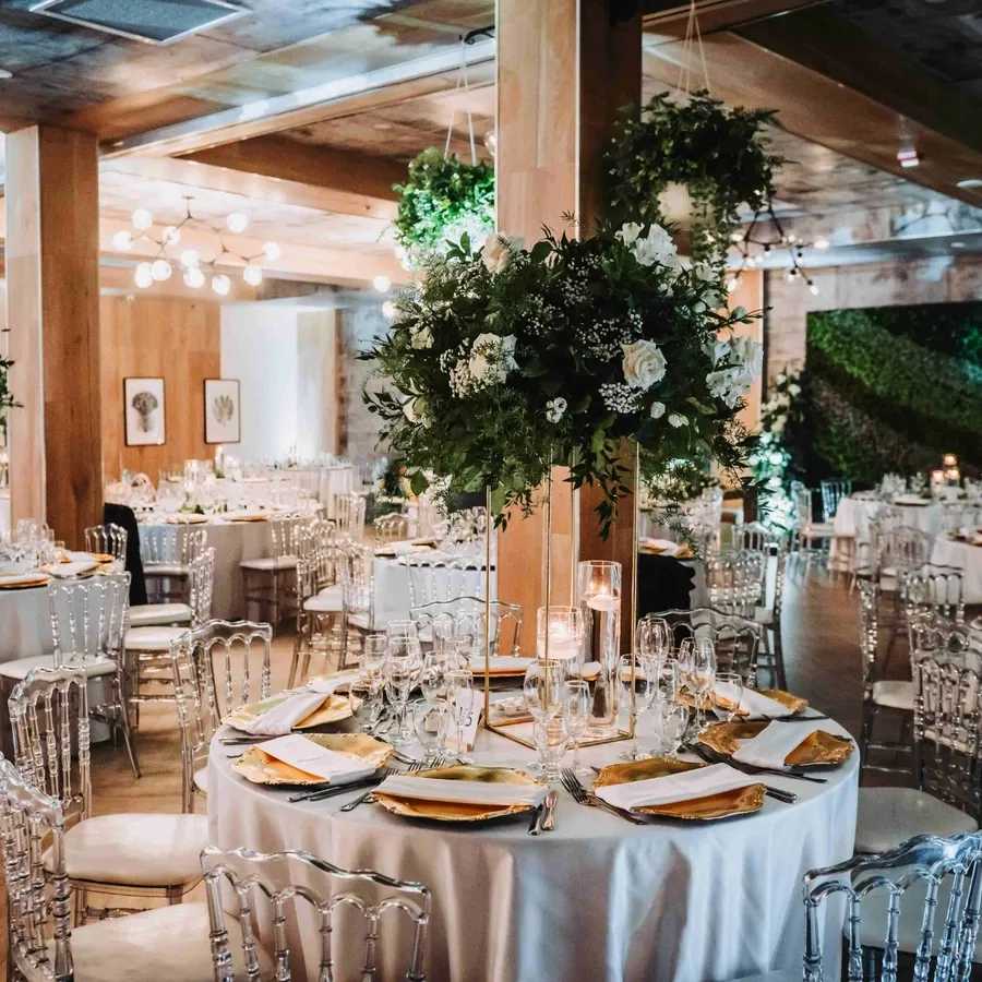 The image showcases Plaza Volare, an exquisite wedding venue featured on Mariage Elegant, Canada. The spacious room is elegantly arranged for a wedding reception, with round tables adorned with white tablecloths, gold-rimmed plates, and crystal glassware. Tall, lush floral centerpieces featuring greenery and white flowers add a touch of sophistication. The room is softly illuminated by numerous candles and overhead chandeliers, creating a warm and inviting ambiance. Clear, modern chairs complement the wooden flooring and walls, contributing to a chic and contemporary aesthetic perfect for a luxurious wedding celebration.