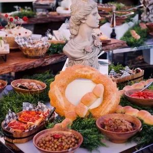 L'image montre une table magnifiquement présentée chez Plaza Volare, une salle de réception spécialisée dans les mariages. Sur la table, divers plats gastronomiques sont disposés, notamment des assiettes de charcuteries, des olives, des pains artisanaux, et des salades appétissantes, créant une atmosphère élégante pour un événement de mariage. Au centre, une statue décorative ajoute une touche artistique, entourée de verdure qui complète la décoration raffinée. Cette présentation met en valeur le souci du détail et l'élégance que Plaza Volare offre comme salle de réception lors de mariages.