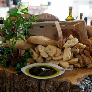 Cette image présente une table élégamment disposée pour une réception de mariage chez le fournisseur Plaza Volare, spécialisé en salles de réception au Canada. Au centre, un assortiment de pains variés, tels que des baguettes et des tranches de pain croustillantes, est joliment arrangé autour d'un panier en bois renversé. Autour, des bouteilles d'huile d'olive apportent une touche méditerranéenne à la scène. Des branches de verdure ornent la table, ajoutant une touche de nature et de sophistication à l'ensemble. Cette présentation culinaire raffinée contribue à créer une atmosphère chaleureuse et accueillante pour les invités du mariage.