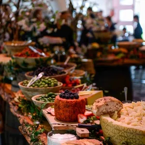 La salle de réception Plaza Volare présente une somptueuse table de buffet ornée pour un mariage. De nombreux plats de charcuterie, fromage, pains, légumes frais et fruits sont disposés élégamment sur des plateaux en bois rustiques. Les décorations florales et feuillues ajoutent une touche naturelle et festive à l'ensemble. L'éclairage doux crée une ambiance chaleureuse, parfaite pour une célébration de mariage.