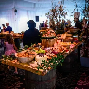 La salle de réception Plaza Volare présente une magnifique disposition de tables en bois rustiques ornées de divers plateaux de mets délicats et fromages, entourés de noix, fruits frais et verdure. L'élégant décor floral et les branches torsadées ajoutent une touche naturelle et enchantée à l'atmosphère, idéale pour un mariage. En arrière-plan, on peut apercevoir des invités admirant l'agencement sous un plafond drapé de tissu blanc, créant une ambiance accueillante et festive.