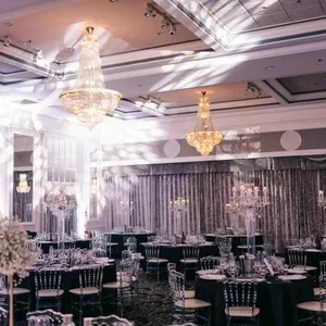 La salle de réception Plaza Volare se présente dans une ambiance élégante et raffinée, idéale pour un mariage. Le plafond est orné de chandeliers somptueux qui projettent des lumières scintillantes dans toute la pièce. Des tables rondes, soigneusement dressées avec des nappes blanches et des chaises transparentes, ajoutent une touche de modernité. Les centres de table sont magnifiquement décorés de bouquets floraux et de bougies, créant une atmosphère romantique. Les tentures murales luxueuses et les hauts plafonds augmentent le charme sophistiqué de cette salle de mariage exceptionnelle au Canada.
