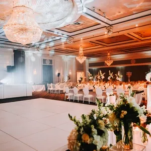 La salle de réception Plaza Volare est magnifiquement décorée pour un mariage. On y voit de grandes tables rondes ornées de centres de table floraux élégants et de chandeliers scintillants suspendus au plafond. La salle est spacieuse, avec une piste de danse centrale entourée de chaises blanches assorties. Un éclairage doux et chaleureux crée une ambiance romantique parfaite pour un mariage.