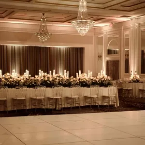La salle de réception Plaza Volare, présentée ici dans un cadre élégant pour un mariage, est ornée de chandeliers étincelants suspendus au plafond, ajoutant une touche de glamour à l'ambiance. Une longue table de banquet est décorée de magnifiques arrangements floraux blancs et de bougies allumées, créant une atmosphère romantique et sophistiquée. Les murs sont ornés de panneaux de bois blanc, et les rideaux élégants encadrent la scène, offrant un cadre parfait pour une célébration de mariage mémorable.