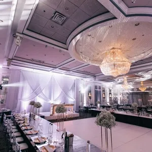 La photo montre la somptueuse salle de réception Plaza Volare, idéale pour un mariage élégant. La pièce est magnifiquement décorée, avec un grand lustre scintillant suspendu au plafond, conférant une atmosphère luxueuse. Les tables sont élégamment dressées avec des nappes blanches et ornées de centres de table floraux. Un espace de danse spacieux est situé au centre, entouré de chaises disposées pour accueillir les invités. L'éclairage subtil et les drapés en tissu ajoutent une touche de raffinement, parfait pour célébrer un événement mémorable.