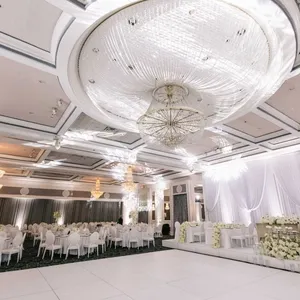 La photo présente une élégante salle de réception proposée par le prestataire Plaza Volare, spécialisée dans les mariages. Le plafond est orné de somptueux lustres scintillants, créant une ambiance lumineuse et raffinée. Les tables sont soigneusement disposées avec des nappes blanches et entourées de chaises élégantes. À droite, une scène décorée de rideaux blancs et de fleurs, idéale pour les cérémonies ou discours durant un mariage, est visible. Cette image illustre parfaitement l’atmosphère sophistiquée que Plaza Volare offre aux couples pour leur événement spécial.