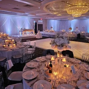 La salle de réception Plaza Volare est magnifiquement aménagée pour un mariage élégant. De grandes tables rondes sont dressées avec des nappes blanches, des assiettes en porcelaine blanche, des verres à vin étincelants et des couverts brillants. Chaque table est ornée d'un centre de table floral avec des fleurs blanches, placé au milieu de bougies votives qui créent une ambiance chaleureuse et romantique. En arrière-plan, de grandes tentures blanches recouvrent les murs, rehaussées par un éclairage discret qui ajoute une teinte douce à la pièce. Un grand lustre en cristal accroché au plafond illumine la piste de danse, prête à accueillir les invités. L’ensemble incarne un cadre raffiné et accueillant, idéal pour célébrer un mariage.