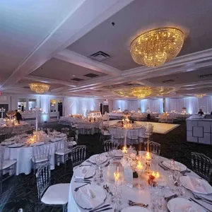 Cette image montre l'intérieur élégant de la salle de réception Plaza Volare, un lieu de mariage proposé par Mariage Elegant au Canada. La salle est magnifiquement décorée avec de somptueux lustres dorés éclairant l'espace. Les tables rondes sont dressées avec des nappes blanches, des verres à vin, de la vaisselle fine et des bougies allumées, créant une ambiance chaleureuse et romantique. Les chaises transparentes ajoutent une touche de modernité à l'ensemble. L'éclairage subtil et les draperies blanches aux murs renforcent l'atmosphère sophistiquée, idéale pour un mariage raffiné.