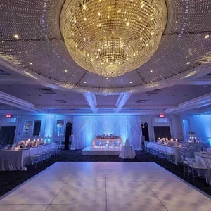 Cette image montre une somptueuse salle de réception, Plaza Volare, idéale pour les mariages. Le plafond est orné d'un lustre cristallin étincelant. Les tables, élégamment drapées de nappes blanches, sont alignées de chaque côté et décorées de bougies scintillantes. Au centre, une piste de danse spacieuse se trouve devant une table d'honneur, ornée de fleurs délicates. L'éclairage bleu doux crée une ambiance romantique et sophistiquée, parfaite pour une célébration de mariage mémorable.