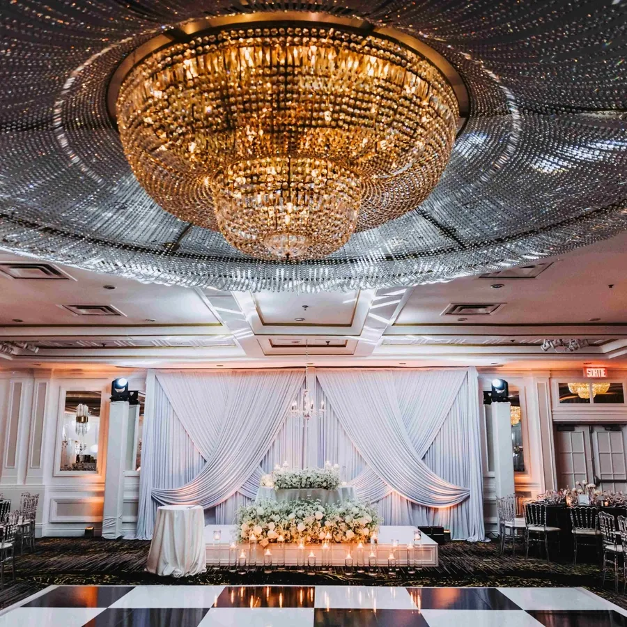 The image displays a luxurious wedding venue at Plaza Volare, categorized under Wedding Venues for Mariage Elegant, Canada. The grand ballroom features an opulent crystal chandelier as its centerpiece, casting a warm glow over the setting. Elegant drapery cascades behind a beautifully adorned sweetheart table, which is embellished with white floral arrangements. The room is set with polished tables, each elegantly dressed with white tablecloths and surrounded by silver Chiavari chairs. Soft, ambient lighting and decorative molding add sophistication, making it an ideal setting for a wedding celebration.