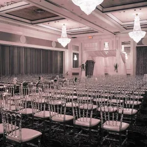 La salle de réception Plaza Volare, spécialisée dans les mariages, est magnifiquement aménagée pour une cérémonie. L'image montre une grande salle élégamment décorée, avec des chaises transparentes soigneusement alignées sur un sol à motifs complexes. De somptueux lustres en cristal pendent du plafond, apportant une lumière douce et chaleureuse à l'espace. Une arche décorative blanche, ornée de fleurs, est située à l'avant, créant un point focal parfait pour les mariages. Les murs présentent des détails architecturaux raffinés qui ajoutent à l'ambiance sophistiquée de la salle.