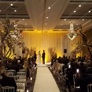 Dans cette image de Plaza Volare, une salle de réception spécialisée dans les mariages, une cérémonie élégante est en cours. La pièce est magnifiquement décorée avec des luminaires en cristal suspendus au plafond et des colonnes de rideaux dorés derrière l'autel. Des chaises blanches disposées en rangées accueillent les invités, tandis que des branches florales ornent les allées, ajoutant une touche naturelle et romantique. Une allée centrale mène à un autel où deux personnes échangent leurs vœux, dans une ambiance chaleureuse et lumineuse. Cette scène illustre parfaitement le cadre prestigieux et sophistiqué que Plaza Volare propose pour les mariages au Canada.