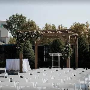 Dans cette image de Mariage Elegant, découvrez la salle de réception Plaza Volare, une magnifique installation extérieure idéale pour les mariages. On y voit une pergola en bois élégamment décorée de roses blanches et roses, et entourée de verdure luxuriante. Des rangées de chaises blanches sont disposées de manière ordonnée, prêtes à accueillir les invités. Un pupitre transparent est placé sous la pergola, parfait pour les cérémonies. Cela crée une ambiance romantique et sophistiquée pour un mariage en plein air.