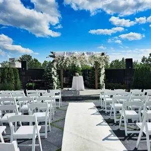 Cette image montre une élégante salle de réception en plein air chez Plaza Volare, spécialisée dans les mariages. Une allée blanche mène à une arche magnifiquement décorée de fleurs blanches et de feuillage, idéale pour une cérémonie de mariage. Les chaises blanches sont disposées en rangées de chaque côté de l'allée, prêtes à accueillir les invités. Le ciel bleu avec des nuages épars ajoute une touche de sérénité à cette scène de fête nuptiale.