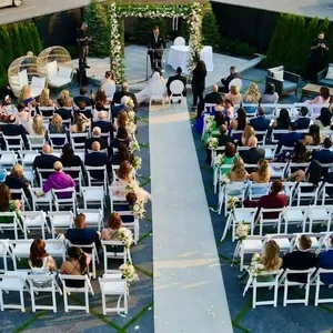 L'image montre un magnifique espace de cérémonie de mariage en plein air à Plaza Volare, une salle de réception réputée. De nombreuses rangées de chaises blanches sont disposées pour accueillir les invités, et une allée centrale mène à une arche élégamment décorée de fleurs blanches et de verdure. Au fond, un couple est debout devant l'arche, probablement en train d'échanger leurs vœux. Deux grands cœurs en osier sont visibles sur la gauche, ajoutant une touche romantique à l'ensemble. Des invités bien habillés sont assis, assistant attentivement à la cérémonie. L'emplacement est entouré de haies vertes, offrant une intimité et un cadre naturel charmant, idéal pour une célébration de mariage.
