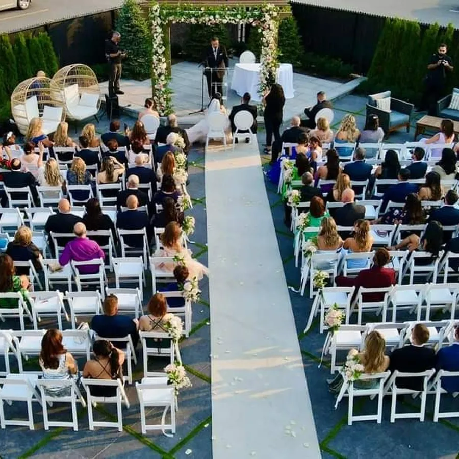 The image showcases a beautifully arranged outdoor wedding ceremony at Plaza Volare, a featured wedding venue on Mariage Elegant, Canada. Guests are seated on white chairs arranged in a semi-circular pattern, facing a decorative arch adorned with lush greenery and white flowers. The aisle is elegantly lined with floral arrangements, adding to the romantic ambiance. In the foreground, a pair of heart-shaped decorations are visible, enhancing the venue