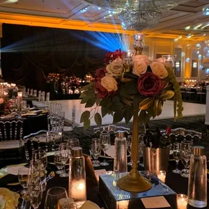 L'image montre une magnifique salle de réception appartenant à Plaza Volare, un lieu populaire pour les mariages au Canada. Au centre, une grande table est magnifiquement décorée avec un grand centre de table floral composé de roses blanches et bordeaux, créant une ambiance romantique. Des chandelles allumées entourent la décoration florale, ajoutant une lueur chaleureuse et élégante. Le plafond est orné de grands lustres scintillants et les lumières tamisées projettent des motifs artistiques sur le plafond, rehaussant l'atmosphère festive de la salle. Des chaises noires avec des accoudoirs transparents entourent les tables, et une vaisselle raffinée est soigneusement placée pour les invités. L'ensemble crée une atmosphère sophistiquée et accueillante, idéale pour célébrer un mariage.