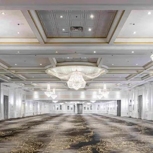 La salle de réception de Plaza Volare, située au Canada, présente un intérieur élégant et spacieux idéal pour les mariages. Le plafond est orné de dorures raffinées et de grands lustres en cristal qui illuminent la pièce avec sophistication. Le sol est recouvert d'une moquette aux motifs complexes, ajoutant une touche de luxe. Les murs blancs et les colonnes encadrent l'espace, permettant une flexibilité dans la décoration. Cette salle de réception offre un cadre prestigieux pour accueillir divers événements nuptiaux, assurant une expérience mémorable pour les invités.