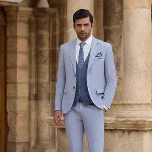 Un homme élégant porte un smoking de mariage moderne créé par le vendeur Baggi. Le costume est de couleur gris clair, accompagné d'un gilet bleu foncé et d'une cravate assortie, parfait pour une cérémonie de mariage raffinée. Le décor en arrière-plan présente des colonnes architecturales classiques en pierre beige, ce qui ajoute une touche sophistiquée et intemporelle à l'image. Ce style est idéal pour les futurs mariés recherchant une tenue chic et contemporaine pour leur grand jour.