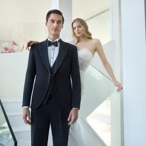 Un homme portant un élégant smoking de mariage de la marque Baggi se tient fièrement au premier plan. Son costume bien taillé est accompagné d'un nœud papillon classique et de détails raffinés. À l'arrière-plan, une femme en robe de mariée ornée de dentelle se tient sur un escalier moderne en verre, posant avec grâce. L'image illustre le style sophistiqué et contemporain des tenues de mariage disponibles chez ce fournisseur, Baggi, dans le cadre des préparatifs de mariages élégants au Canada.