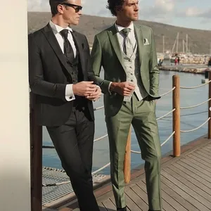 Deux hommes se tiennent côte à côte sur une promenade en bois près de l'eau, élégamment vêtus de smokings de mariage de Baggi. L'un porte un smoking noir classique avec une cravate noire et des chaussures noires brillantes, complété par des lunettes de soleil. L'autre est dans un élégant smoking vert olive avec un gilet gris et une cravate assortie. La scène est éclairée par une lumière naturelle mettant en valeur leur style raffiné, idéal pour une cérémonie de mariage en plein air. À l'arrière-plan, on aperçoit des voiliers et des collines sous un ciel légèrement nuageux, ajoutant une touche de romantisme à l'atmosphère. Cette image représente parfaitement la catégorie de smoking de mariage sur le site Mariage Elegant au Canada.