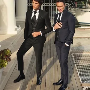 Deux hommes élégamment vêtus posent sur une terrasse en bois devant une balustrade blanche. Le premier homme, à gauche, porte un smoking noir raffiné avec une cravate et un mouchoir de poche assortis, tandis que le second homme, à droite, est vêtu d'un smoking bleu marine avec une cravate noire. Les smokings, proposés par le vendeur Baggi, illustrent la catégorie 'Smoking de mariage' sur Mariage Elegant, un site canadien dédié à la planification de mariages et à la mise en relation avec des prestataires. L'arrière-plan met en valeur un cadre chic et sophistiqué, parfait pour un mariage luxueux.