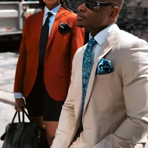 L'image montre un homme et une femme en tenue formelle, idéalement adaptée pour un mariage. L'homme porte un smoking élégant beige de la catégorie 'Smoking de mariage', accompagné d'une chemise blanche et d'une cravate à motifs bleus. Une pochette assortie orne la poche de son smoking. La montre dorée ajoute une touche de luxe. À ses côtés, la femme est habillée d'une veste rouge sophistiquée avec une chemise blanche, une cravate noire, et un short noir. Elle tient un sac à main noir, et ses lunettes de soleil complètent son look chic. Cette image illustre le style moderne et raffiné offert par le vendeur Baggi, idéal pour un mariage contemporain organisé par Mariage Elegant au Canada.