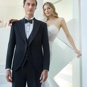 Un couple élégant pose dans un cadre moderne et lumineux. L'homme porte un smoking de mariage raffiné de Baggi, avec une veste noire ornée de détails scintillants et un nœud papillon classique. La femme à ses côtés est vêtue d'une somptueuse robe de mariée blanche avec un décolleté en cœur et des détails en dentelle. Cet ensemble illustre l'élégance et le style pour des cérémonies de mariage sophistiquées, en harmonie avec le thème du site Mariage Elegant au Canada.