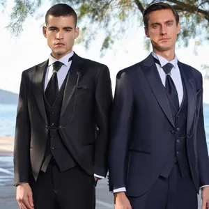 Deux hommes posent élégamment sous les arbres près d'un bord de mer. Ils portent des smokings de mariage raffinés de la boutique Baggi, spécialisés dans les tenues de mariage. Le mannequin de gauche arbore un smoking noir avec un gilet assorti, une chemise blanche et une cravate noire. À droite, le mannequin porte un smoking bleu foncé avec des détails similaires. Ces tenues de cérémonie, idéales pour un mariage élégant, mettent en valeur la qualité et le savoir-faire de la marque Baggi, spécialisée dans les smokings de mariage pour le site Mariage Elegant, Canada.