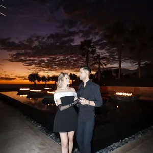 Un couple élégant se tient devant un spectaculaire coucher de soleil, créant une ambiance romantique idéale pour une photo de mariage. La femme porte une robe noire chic tandis que l'homme est habillé élégamment en noir. La scène se déroule près d'une piscine reflétant la lueur du ciel nocturne et des flammes douces illuminent l'arrière-plan. Cette image est capturée par Tiara Studio, experts en photographie de mariage, pour le site Mariage Elegant au Canada.