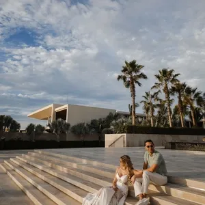 Dans cette image fournie par Tiara Studio, une entreprise spécialisée dans la photographie de mariage, un couple est assis sur de larges marches en pierre sous un ciel partiellement nuageux. La mariée porte une robe blanche élégante et le marié est vêtu de manière décontractée avec une chemise verte et un pantalon clair. Ils sont entourés de palmiers, et l'architecture moderne de l'arrière-plan ajoute une touche contemporaine à la scène. Cette photo capture un moment romantique et serein, idéal pour illustrer l'expertise de Tiara Studio dans l'immortalisation des moments précieux des mariages.