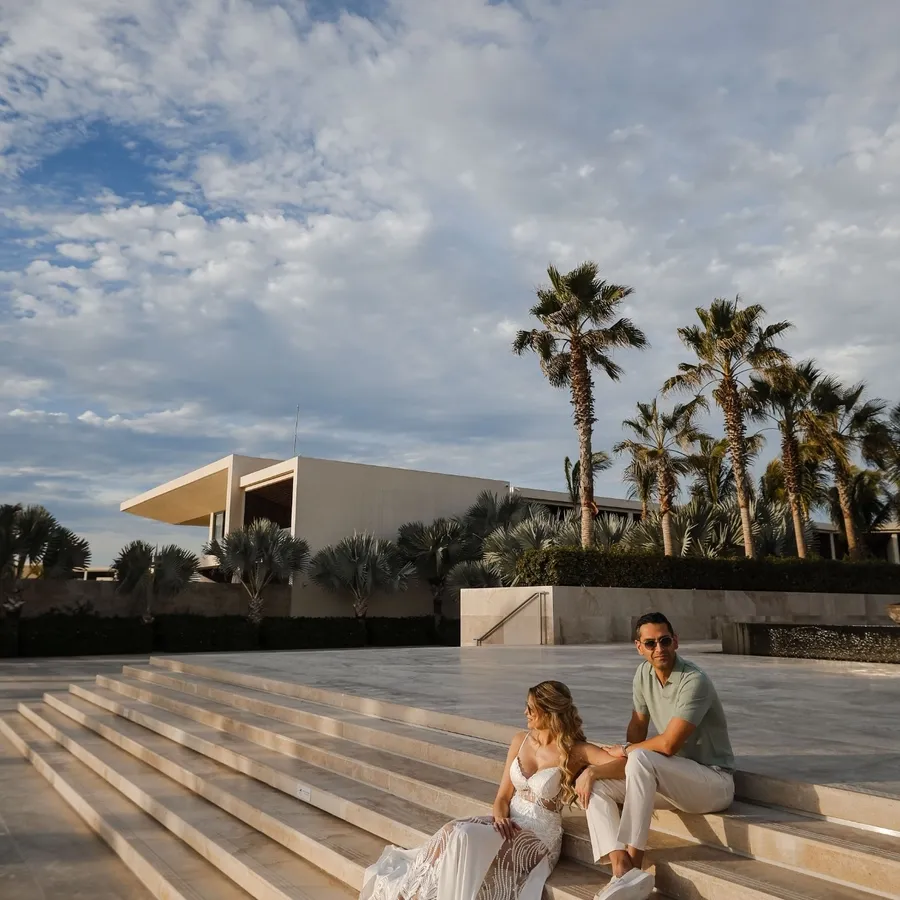 Dans cette image fournie par Tiara Studio, une entreprise spécialisée dans la photographie de mariage, un couple est assis sur de larges marches en pierre sous un ciel partiellement nuageux. La mariée porte une robe blanche élégante et le marié est vêtu de manière décontractée avec une chemise verte et un pantalon clair. Ils sont entourés de palmiers, et l