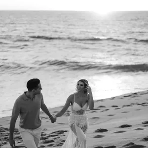 Un couple marche main dans la main sur une plage au coucher du soleil, capturé en noir et blanc par Tiara Studio, spécialisé en photographie de mariage. La mariée porte une robe élégante avec des détails en dentelle, et le marié est vêtu de manière décontractée. L'ambiance romantique de la mer et la lumière douce du soleil couchant ajoutent une touche magique à ce moment partagé, illustrant l'art de Tiara Studio dans l'immortalisation des souvenirs précieux des mariages.
