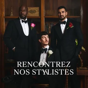 Trois hommes posent élégamment dans des smokings de mariage noirs, chacun portant une chemise blanche et un nœud papillon noir. Des roses colorées ornant leurs boutonnières ajoutent une touche de sophistication. Placés devant une bibliothèque en bois, ils représentent le style raffiné de Surmesur, un prestataire de tenues pour mariages.