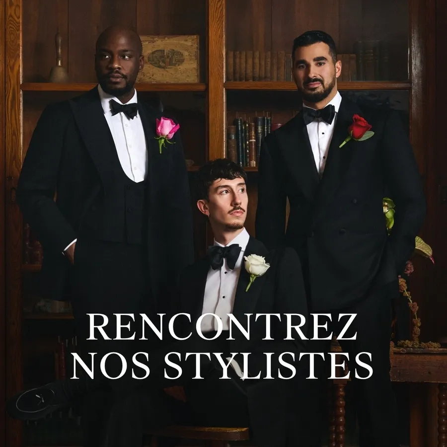 Trois hommes posent élégamment dans des smokings de mariage noirs, chacun portant une chemise blanche et un nœud papillon noir. Des roses colorées ornant leurs boutonnières ajoutent une touche de sophistication. Placés devant une bibliothèque en bois, ils représentent le style raffiné de Surmesur, un prestataire de tenues pour mariages.