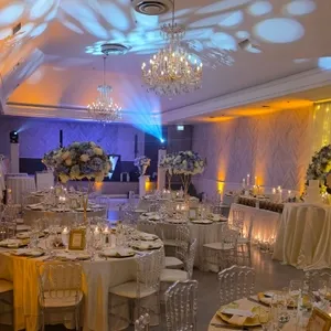 La salle de réception de The PMG Group, sous la catégorie 'Salle de réception', présente un décor élégant et raffiné pour un mariage. La pièce est ornée de superbes lustres en cristal suspendus au plafond, offrant un éclairage brillant et chaleureux. Les tables rondes sont élégamment dressées avec des nappes blanches, des couverts en argent et des bouquets de fleurs blanches et bleues en centres de table. Des chaises transparentes ajoutent une touche moderne au décor classique. L'architecture de la salle, avec ses murs ornés de motifs subtils et un éclairage d'ambiance coloré, crée une atmosphère romantique idéale pour célébrer un mariage.