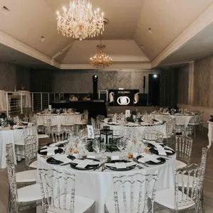 La photo montre une élégante salle de réception préparée pour un mariage, fournie par The PMG Group, dans la catégorie salle de réception. La pièce est ornée de lustres scintillants qui ajoutent une touche de glamour. Les tables rondes sont magnifiquement dressées avec des nappes blanches impeccables, des couverts raffinés, et des chaises transparentes modernes. Chaque table est centrée avec des bougies et des compositions florales délicates. La scène en arrière-plan est prête pour les discours et les présentations, créant une ambiance accueillante et festive pour les invités.