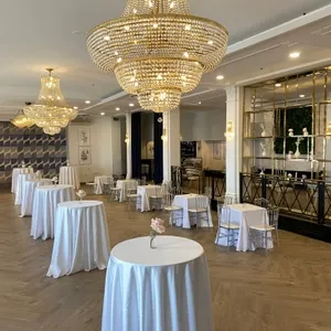 La salle de réception de The PMG Group, présentée ici, est élégamment décorée pour un événement de mariage. De magnifiques lustres en cristal pendent du plafond, diffusant une lumière douce sur la pièce. Des tables rondes, chacune recouverte d'une nappe blanche immaculée et ornée d'un élégant vase avec une fleur, sont disposées de manière espacée sur le parquet en bois clair. Un mur avec un motif géométrique discret et une grande étagère garnie de verdure ajoutent une touche moderne au décor. Cet espace spacieux est idéal pour accueillir des réceptions de mariage au Canada.