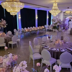 La salle de réception de The PMG Group, un espace de mariage élégant au Canada, est magnifiquement décorée. De grands lustres en cristal illuminent la pièce, créant une atmosphère luxueuse. Les tables rondes sont ornées de nappes élégantes et de magnifiques compositions florales dans des nuances de rose et de violet. Les chaises blanches assorties ajoutent une touche de sophistication. Des rideaux sombres encadrent les fenêtres, et la lumière ambiante contribue à l'ambiance romantique de ce lieu de mariage.