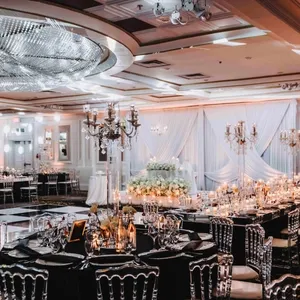 Cette image montre une salle de réception de mariage élégante proposée par The PMG Group dans la catégorie Salle de réception. La pièce est somptueusement décorée avec des tables drapées de nappes noires, entourées de chaises transparentes qui ajoutent une touche moderne. De grands chandeliers en cristal illuminent l'espace, reflétant une lumière douce et romantique. Le plafond est orné d'un vastes lustre en cristal, créant une atmosphère luxueuse. Un fond de drapés blancs et de fleurs blanches ajoute une élégance supplémentaire à l'environnement, ce qui est idéal pour un mariage chic et sophistiqué.