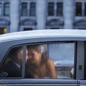 Une mariée et un marié sont assis à l'arrière d'une voiture élégante, probablement une limousine ancienne, sous la pluie. La mariée porte une robe blanche et un diadème étincelant tandis qu'ils se regardent tendrement. À travers la vitre, des gouttes de pluie ajoutent une atmosphère romantique et intime à ce moment capturé. En arrière-plan, un bâtiment historique flou se dresse, accentuant le cadre urbain et sophistiqué. Cette image est un exemple du travail raffiné d'Inhouse Studios Photography dans la catégorie Photographie de mariage pour Mariage Elegant au Canada.