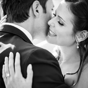 Dans cette image en noir et blanc, capturée par Inhouse Studios Photography pour le site Mariage Elegant au Canada, un couple de mariés échange un moment intime. La mariée sourit tendrement tandis que le marié l'enlace et lui murmure à l'oreille. Elle porte des boucles d'oreilles élégantes et une robe de mariée. Le focus est sur l'émotion et la complicité de cet instant précieux, mettant en valeur l'expertise de la photographie de mariage.