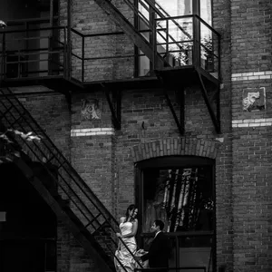 Mariée et marié photographiés sur un escalier en métal contre un mur de briques, donnant un aspect industriel et urbain. La mariée porte une élégante robe blanche, tandis que le marié est en costume sombre. La lumière joue subtilement sur les textures des briques et des éléments métalliques, créant une ambiance romantique et contemporaine. Ce moment capturé par Inhouse Studios Photography est un bel exemple de photographie de mariage dans un cadre unique, mettant en valeur le talent du photographe à immortaliser des instants intimes dans un décor authentique. Catégorie : Photographie de mariage.