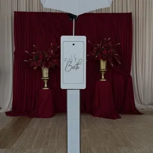 L'image présente un élégant photo booth de la marque 'Le Fotobooth', appartenant à la catégorie des cabines photo, idéal pour les événements de mariage. Le dispositif moderne est surmonté d'un grand parapluie blanc, assurant un bon éclairage. En arrière-plan, des rideaux de velours rouge bordeaux encadrent deux urnes dorées remplies de bouquets de fleurs rouges, ajoutant une touche sophistiquée et festive, typique des décorations de mariage.