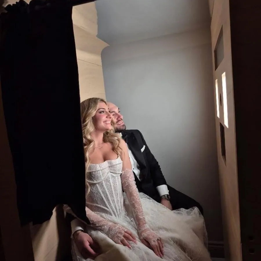 A couple sits inside a modern photo booth provided by Le Fotobooth, categorized under Photo Booth services for weddings. The bride, in an elegant, lace wedding dress with long sleeves, leans affectionately against the groom, who is dressed in a classic black tuxedo. They are both smiling and posing as the booth captures their joyful moment. The ambiance is intimate, with soft lighting creating a romantic atmosphere, typical for capturing special moments at a wedding reception.