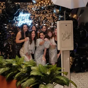 Groupe de femmes souriantes posant devant un photomaton moderne de Le Fotobooth, dans la catégorie des cabines photo pour Mariage Elegant, Canada. Elles sont habillées de robes élégantes et scintillantes, parfaites pour une célébration de mariage. En arrière-plan, un mur de ballons noirs et dorés accompagné d'un panneau lumineux affichant 'Happy Birthday' crée une ambiance festive et sophistiquée. Au premier plan, des plantes vertes ajoutent une touche de fraîcheur à la scène.