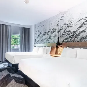 La photo montre une élégante chambre moderne du Doubletree par Hilton Québec Resort, classée dans la catégorie salle de réception pour mariages sur Mariage Elegant, Canada. La pièce dispose de deux grands lits aux draps blancs immaculés, idéals pour accueillir des invités lors d'une célébration de mariage. Un grand mur décoré d'une fresque artistique représentant une scène naturelle ajoute une touche raffinée. À gauche, une petite table avec une machine à café est visible, offrant du confort aux invités. Les rideaux gris encadrent une fenêtre laissant entrer la lumière naturelle, améliorant l'ambiance accueillante de l'espace.